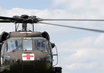 A military medical evacuation (MEDEVAC) helicopter, identified by the prominent Red Cross emblem on its nose, is positioned on a grassy hillside with wildflowers. The aircraft bears the number '745' and appears to be an olive drab UH-60 Black Hawk variant configured for medical operations. The rotors are spinning, suggesting the aircraft is either preparing for takeoff or has just landed.
