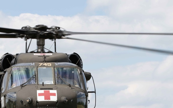 A military medical evacuation (MEDEVAC) helicopter, identified by the prominent Red Cross emblem on its nose, is positioned on a grassy hillside with wildflowers. The aircraft bears the number '745' and appears to be an olive drab UH-60 Black Hawk variant configured for medical operations. The rotors are spinning, suggesting the aircraft is either preparing for takeoff or has just landed.