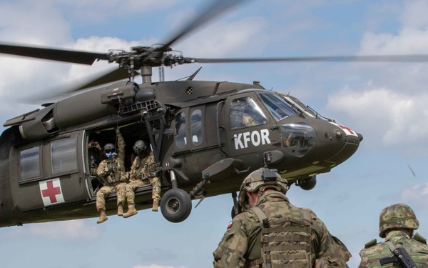 A military helicopter marked 'KFOR' (Kosovo Force) with a Red Cross medical insignia hovers low over a group of ground soldiers in camouflage uniforms. The helicopter appears to be a UH-60 Black Hawk operated by the U.S. Army, with crew members visible in the open side door. Ground troops, carrying rifles and tactical gear, observe the helicopter during what appears to be a training exercise or operational demonstration.