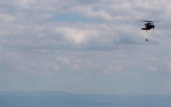 A military helicopter, appearing to be a UH-60 Black Hawk, is visible in the upper right portion of the image with a person suspended below it on a hoist or rope, suggesting a rescue or rappelling operation. The scene is set over a expansive green, hilly landscape with forested terrain in the foreground and a wide valley visible in the background. The sky is partly cloudy with good visibility across the terrain.