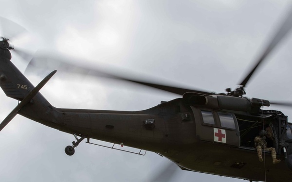 A military medevac helicopter, marked with a red cross and 'KFOR' designation, is airborne with a soldier being hoisted or rappelling on a rope beneath it. At least one additional crew member is visible in the open side door of the aircraft. The scene appears to depict a hoist rescue or insertion training exercise.