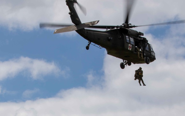 A military medical evacuation (MEDEVAC) exercise is underway, featuring a UH-60 Black Hawk helicopter marked with a Red Cross symbol hovering above a grassy field. A soldier is being hoisted or rappelling on a cable beneath the aircraft. On the ground, two groups of soldiers in different camouflage patterns observe the operation.