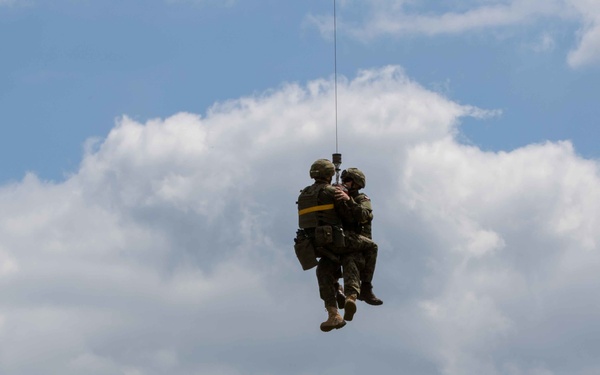 A military hoist/extraction operation is underway in an open grassy field. Two personnel are suspended in the air on a hoist line, with one appearing to hold or support the other, suggesting a casualty evacuation or MEDEVAC training exercise. A third soldier on the ground is gesturing upward, likely directing or signaling to the aircraft above.