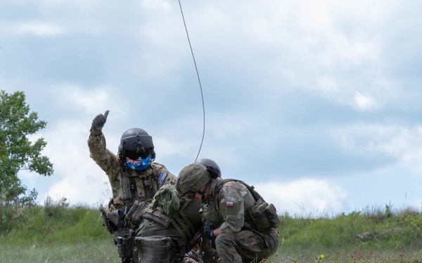 Two military personnel in combat gear are crouched on a grassy hillside, with one soldier raising a hand upward toward a cable or wire extending into the sky. Both wear helmets and tactical vests, and visible flag patches suggest multinational participation, likely in a joint military exercise. The setting appears to be an open field training environment under overcast skies.