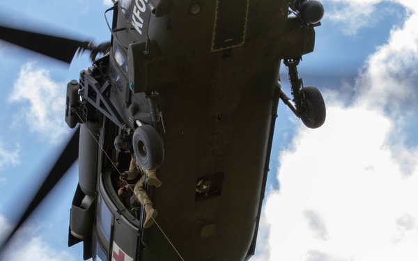 A military medical evacuation (MEDEVAC) helicopter, identifiable by the red cross marking on its fuselage, is conducting a hoist rescue operation in flight. Two personnel are suspended below the aircraft on a hoist cable, with one appearing to carry or support another individual. A crew member is visible leaning out of the open side door managing the operation.