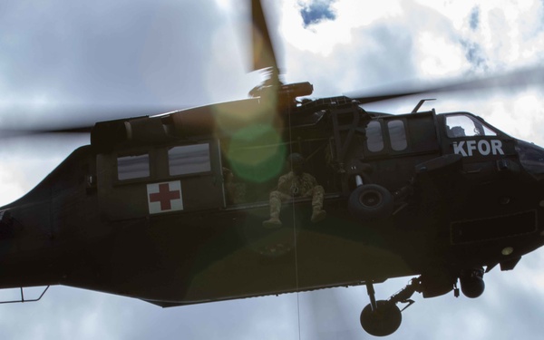 A military medical evacuation (MEDEVAC) helicopter marked with a Red Cross and 'KFOR' designation is airborne against a partly cloudy sky. Two personnel are suspended below the helicopter on a hoist cable, appearing to conduct a rescue or extraction operation. A crew member is visible seated in the open side door of the aircraft.