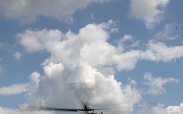 A military medical evacuation (MEDEVAC) helicopter, marked with a Red Cross and 'KFOR' designation, hovers low over a grassy field while several soldiers in MultiCam/OCP camouflage uniforms observe from the ground. The scene appears to be a MEDEVAC training exercise or actual casualty evacuation operation conducted under NATO's Kosovo Force (KFOR) mission. The helicopter is a UH-60 Black Hawk variant configured for medical transport.