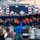 A patriotic outdoor concert is taking place in a stadium setting, featuring a large choir dressed in navy blue robes and a military band in dress uniforms performing on a decorated stage. A conductor stands with his back to the audience, directing the ensemble in front of a large LED screen displaying stars and stripes imagery. An audience seated in white folding chairs watches the performance, with some members recording on phones.