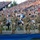 Four military personnel in U.S. Army OCP (Operational Camouflage Pattern) uniforms are seated on a raised platform/stage at what appears to be a large outdoor stadium event. They are smiling and appear to be honored guests at a public ceremony, likely a patriotic or university event given the visible BYU logo in the background. The stadium is filled with a large crowd of spectators.