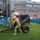 Military personnel in camouflage uniforms are operating a ceremonial artillery cannon on a grass field during a large public event at a stadium. A choir dressed in dark blue robes stands nearby facing a massive crowd filling the stadium stands. The event appears to be a patriotic celebration, likely a Fourth of July or military appreciation ceremony, with patriotic banners and stage equipment visible.