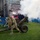 Two U.S. Army soldiers in camouflage uniforms are operating a ceremonial artillery cannon on a grass field during what appears to be a large public event or celebration. The cannon has just been fired, as evidenced by the white smoke billowing from the barrel. A large crowd fills the stadium stands in the background, and a choir in dark robes is visible near a performance stage.