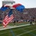 A parachutist is landing on a football field while carrying a large American flag, as part of a pre-game or halftime ceremony at a packed stadium. The red, white, and blue parachute is still inflated as the jumper touches down on the turf. Several individuals in military uniforms and civilian clothing are running toward the landing zone to assist.