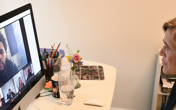 A woman with long red hair is seated at a white desk participating in a video conference call on an Apple iMac computer. The screen displays multiple participants in a grid layout typical of platforms like Zoom or Microsoft Teams. The setting appears to be a home office or private workspace.