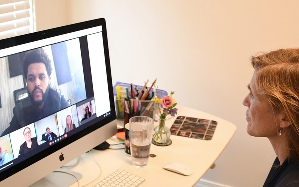 A woman with red hair is participating in a video conference call on an Apple iMac desktop computer at a home office desk. The screen displays multiple participants in a grid layout typical of video conferencing software such as Zoom or Microsoft Teams. The workspace is organized with various desk accessories, documents, and personal items visible.