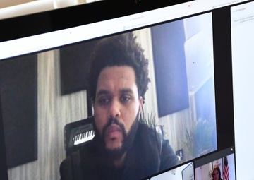 A computer monitor displays an active video conference call, likely on Google Meet, showing multiple participants in a grid layout. The largest tile features a man with natural curly hair and a beard in what appears to be a home or studio setting with a keyboard instrument visible in the background. Several other participants are visible in smaller tiles at the bottom of the screen.