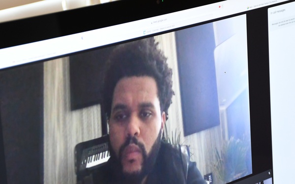 A computer monitor displays an active video conference call, likely on Google Meet, showing multiple participants in a grid layout. The largest tile features a man with natural curly hair and a beard in what appears to be a home or studio setting with a keyboard instrument visible in the background. Several other participants are visible in smaller tiles at the bottom of the screen.