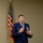 A senior U.S. Air Force officer in dress blue uniform is speaking at a formal event or briefing, holding a microphone. He is positioned in front of an American flag against a neutral beige wall. His uniform displays numerous service ribbons, badges, and rank insignia indicating a high-ranking officer.