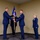 A formal U.S. Air Force change of command ceremony is taking place indoors. Two officers in dress blue uniforms are exchanging a unit guidon (flag on a staff), which is the traditional centerpiece of such ceremonies. A female officer stands at attention to the right, and a flag bearer holds position in the background.