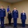 A formal U.S. Air Force military ceremony featuring four personnel in dress blue uniforms standing at attention. Three officers are positioned in the foreground while a non-commissioned officer holds a unit guidon flag in the background. The setting includes the U.S. flag, an Arkansas state flag, and a blue Air Force flag, suggesting a change of command or similar official ceremony.
