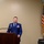 A United States Air Force officer in dress blue uniform stands at a wooden podium, apparently delivering a speech or address at an official ceremony. The setting appears to be an indoor venue with an American flag displayed on a stand to the right. The officer's uniform displays multiple service ribbons, wings, and rank insignia consistent with a senior officer.