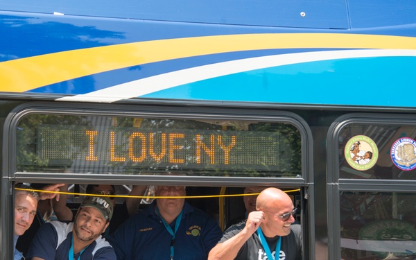 A group of people are leaning out of the windows of an MTA Electric Bus, smiling and gesturing enthusiastically. The bus displays 'I LOVE NY' on its LED destination sign, and the lower body of the bus reads 'ELECTRIC BUS'. The scene appears to be a public event or promotional occasion, with participants wearing lanyards and branded apparel.