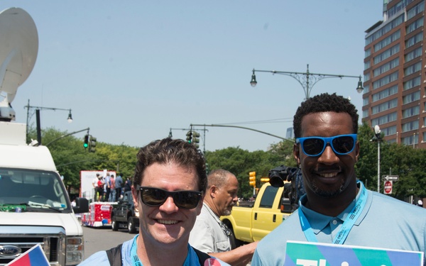 Two individuals wearing light blue NYC Health t-shirts and lanyards pose together at an outdoor public health event in New York City. They are holding vaccination campaign signs reading 'New Vaxx City' and 'This is our SHOT NYC'. The setting appears to be a street or public plaza with media satellite trucks, traffic signals, and a tall building visible in the background.