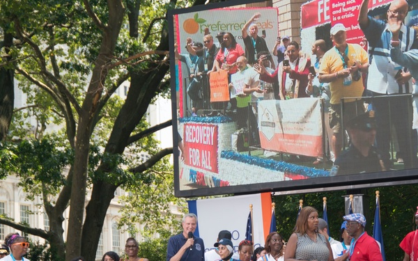 A public outdoor event or rally is taking place on a stage, with a tall man speaking into a microphone at the center surrounded by a diverse group of people. A large LED screen behind the stage displays a live feed of the event, showing banners including 'Recovery For All' and 'Preferred Meals.' The setting appears to be a civic plaza or park, with trees and a classical building visible in the background.