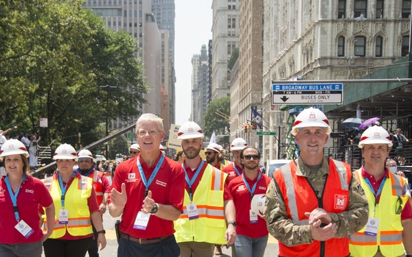 A group of individuals wearing white hard hats, red shirts, and high-visibility yellow/orange vests are marching down Broadway in New York City during what appears to be a ticker-tape parade. The group includes both civilians and a military personnel member in camouflage uniform. Confetti and paper streamers litter the street, and spectators line the barricaded sidewalks.