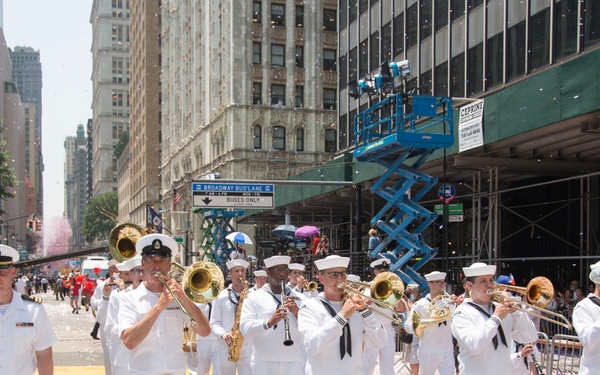 A U.S. Navy marching band in full white dress uniforms parades down a major urban street, identifiable as Broadway in New York City based on visible street signage. The street is covered in confetti and ticker tape, suggesting a celebratory parade event. Spectators line the sidewalks behind metal barricades while a blue scissor lift and construction scaffolding are visible on the right side.