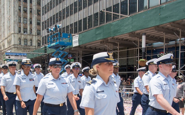 A formation of U.S. Coast Guard personnel in dress uniform marches down a city street covered in ticker tape and confetti, consistent with a ticker-tape parade in New York City. The street sign visible in the background reads 'Broadway' confirming the Manhattan location. Both male and female service members are present, wearing white service dress uniforms with navy trousers and white combination covers.