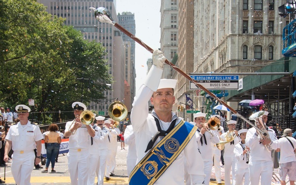A U.S. Navy marching band parades down a major urban street, identifiable as Broadway in New York City based on visible street signage. The drum major leads the formation with a raised baton while band members in white Navy dress uniforms play brass instruments. Confetti covers the street and sidewalks, indicating a celebratory public event.