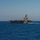 A large US Navy aircraft carrier is underway on open blue ocean under clear skies. The carrier is viewed from a slight angle showing its port side and bow, with aircraft visible on the flight deck and the island superstructure prominently rising on the starboard side. A white wake is visible behind the vessel indicating forward movement at speed.