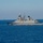 A large grey naval warship is underway at sea, displaying hull numbers on both bow and stern sections. The vessel appears to be a modern destroyer or frigate with sophisticated radar and weapons systems visible on its superstructure. The ship is creating a visible wake as it moves through calm, deep blue open ocean waters.