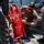 A military or naval personnel member in a brown t-shirt and sunglasses is crouching down to inspect or adjust a red Stokes litter (rescue stretcher) with a metal frame on what appears to be a ship's deck or flight deck. Two other individuals in olive/flight suits are standing nearby. The setting suggests a medical evacuation or search-and-rescue equipment check.