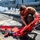A military service member in olive/tan uniform is kneeling on the flight deck of a naval vessel, handling a red rescue/medical litter (stretcher) with restraint straps. A U.S. Navy helicopter is visible in the background, chained to the deck. The setting appears to be an aircraft carrier or amphibious assault ship flight deck at sea.