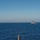 The image shows two naval vessels underway on open blue ocean under a clear sky. A larger vessel, consistent with an aircraft carrier, is visible in the distant left background, while a smaller warship — likely a frigate or destroyer — is more prominently positioned in the right foreground. The scene appears to depict a naval formation or escort operation.