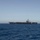 A large aircraft carrier is photographed at sea from a distance, showing its full profile against a clear blue sky. The vessel appears to be underway or stationary in open ocean, with aircraft visible on the flight deck. The image was taken from another vessel or low vantage point at sea level.
