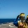 Two naval personnel wearing yellow flight deck vests and helmets with communication headsets stand on a ship's flight deck observing a helicopter performing a vertical replenishment (VERTREP) operation. The helicopter, visible in the upper left, is airborne and carrying a sling-loaded cargo net beneath it. The scene takes place over open ocean under clear blue skies.