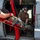 A military or coast guard crew member wearing a flight helmet and brown t-shirt is inside a helicopter, assisting with loading a patient on a red Stokes litter/stretcher through the side door. The scene appears to be a medevac or search-and-rescue operation, with the individual kneeling inside the aircraft cabin to receive the casualty. Equipment including straps, harnesses, and rescue gear are visible throughout the frame.