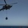 A military helicopter, appearing to be a U.S. Navy MH-60R Seahawk, is conducting a vertical replenishment (VERTREP) operation over open ocean. The helicopter is carrying a cargo net loaded with supplies suspended beneath it on a hoist line. A naval vessel is partially visible in the lower right corner of the frame.