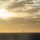 A wide ocean scene photographed at or near sunset, with strong golden light emanating from the upper left. In the distant center of the frame, a large vessel — consistent in silhouette with an aircraft carrier — is visible on the horizon, partially obscured by haze. A second, smaller vessel appears to be nearby.