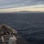 The image shows the bow of a naval warship underway at sea, photographed from an elevated position looking forward. A mountainous coastline is visible on the horizon in the background, along with a distant vessel. The lighting suggests either dawn or dusk conditions with overcast skies and warm horizon tones.