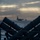 A naval scene photographed from the deck of a warship, showing missile launcher tubes in the foreground arranged in a V-shaped configuration. In the background, at least one large vessel — identifiable by its flat deck profile as an aircraft carrier — is visible on the horizon. The image is taken during overcast, low-light conditions, likely at dusk or dawn, giving the scene a dramatic silhouetted appearance.