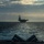 A large aircraft carrier is visible on the horizon, photographed from the deck of another naval vessel at sea. The image is taken during overcast, diffuse lighting conditions, giving the scene a dramatic silhouetted appearance. The foreground shows deck equipment of the observing vessel, while the carrier dominates the mid-distance.