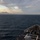 A naval warship is underway at sea during twilight or early morning hours, with a mountainous landmass visible on the distant horizon. The photograph is taken from an elevated position on the ship looking forward over the bow. The sea conditions appear moderate with light chop and whitecaps visible across the water surface.