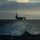 A large aircraft carrier is visible in silhouette on the open ocean, photographed from the deck of another vessel at dusk or dawn. The image captures a dramatic atmospheric scene with diffused light breaking through overcast clouds on the horizon. A smaller vessel is also faintly visible to the left of the carrier in the distance.