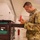 A U.S. Air Force service member in OCP (Operational Camouflage Pattern) uniform is examining a small red and white item, possibly a tagged component or part, while holding paperwork. He is standing next to a black stackable storage/equipment container with green foam inserts and a label reading 'MA02TDYB3'. The setting appears to be an indoor military logistics, supply, or maintenance facility.