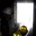 Three individuals wearing hard hats are positioned inside a dark structure facing a brightly lit opening, with water spray or mist visible throughout the scene. Two individuals wear yellow firefighter-style helmets and one wears a white helmet with red markings. The high-contrast lighting and water droplets suggest a firefighting training exercise or emergency response scenario.