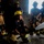 A group of firefighters, likely trainees, are participating in an indoor hose handling exercise inside a concrete structure. One firefighter in the foreground is kneeling and directing a pressurized water stream from a hose nozzle while others observe. The scene appears to be a structured training drill with multiple participants in full turnout gear and yellow helmets.