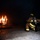 Three firefighters in full protective gear are crouching low and advancing toward a controlled wood pallet fire inside a smoke-filled structure. The scene appears to be a live fire training exercise, with the firefighters holding a charged hose line in a low-crawl position. Smoke fills the upper portion of the enclosed space while flames illuminate the otherwise dark environment.